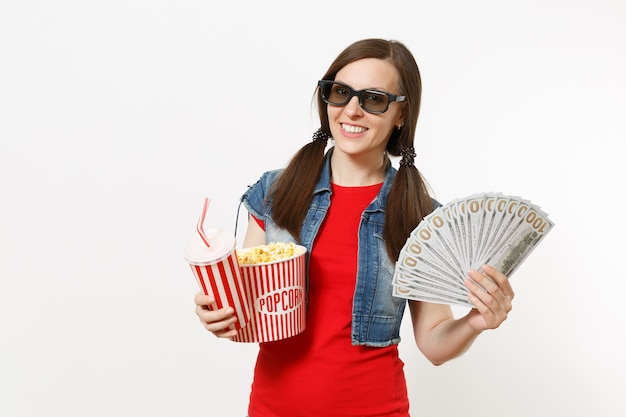 Portrait d'une jeune femme souriante dans des lunettes 3d regardant un film, tenant un seau de pop-corn, une tasse de soda ou de cola et un paquet de dollars, de l'argent liquide isolé sur fond blanc. Les émotions au cinéma.