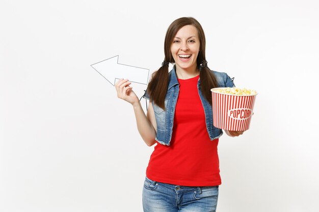 Portrait de jeune femme souriante dans des vêtements décontractés en regardant un film, tenant un seau de pop-corn, pointant la flèche blanche de côté sur fond isolé sur fond blanc. Émotions dans le concept de cinéma.