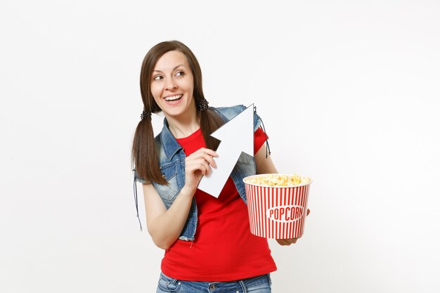 Portrait de jeune femme souriante dans des vêtements décontractés en regardant un film, tenant un seau de pop-corn, pointant la flèche blanche vers le haut sur fond isolé sur fond blanc. Émotions dans le concept de cinéma.