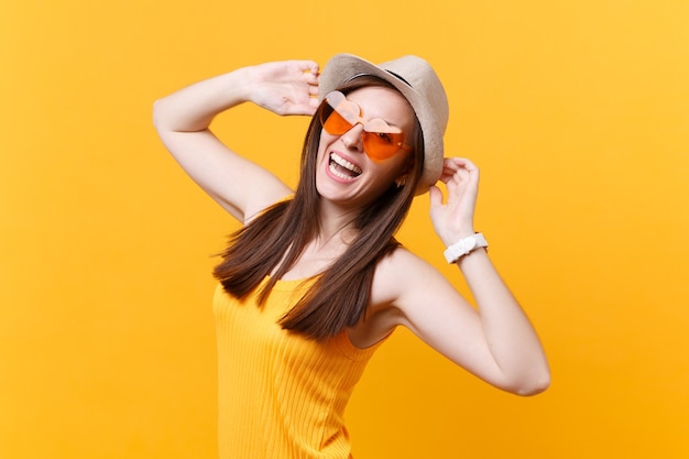 Portrait d'une jeune femme souriante excitée au chapeau d'été de paille, des lunettes orange mettent les mains sur la tête, regarde de côté isolé sur fond jaune. Les gens émotions sincères, concept de style de vie. Espace publicitaire.