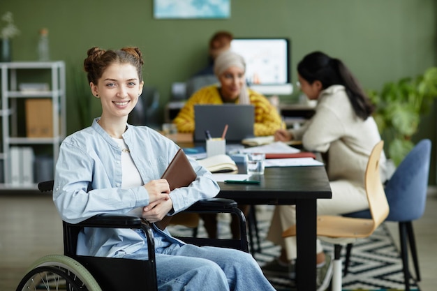 Portrait d'une jeune femme souriante en fauteuil roulant regardant la caméra tout en travaillant au bureau avec divers