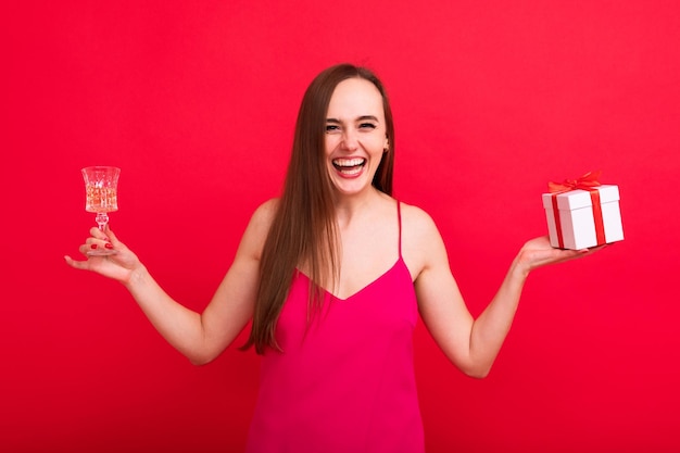 Portrait d'une jeune femme souriante heureuse dans une robe élégante rose tenant un verre de champagne et une boîte avec un cadeauFête de vacances