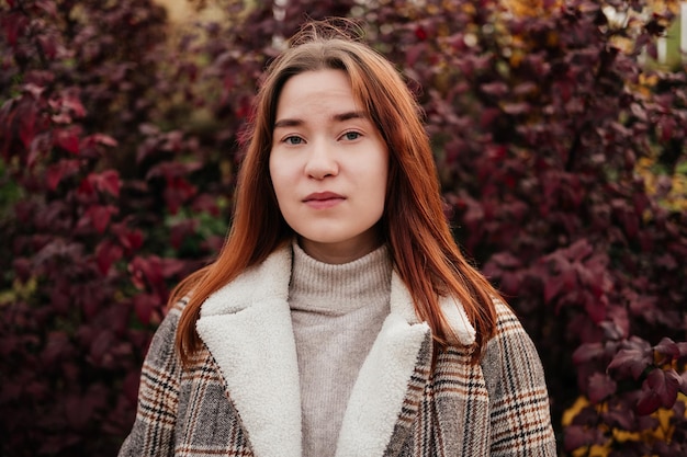 Portrait d'une jeune femme souriante en manteau et pull debout devant un buisson de feuilles rouges et regardant la caméra