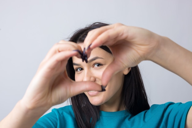 Portrait d'une jeune femme souriante montrant un geste cardiaque avec ses doigts isolés sur gris