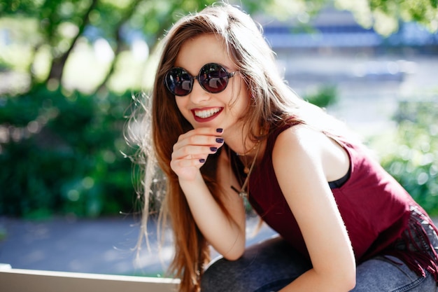 Portrait d'une jeune femme souriante portant des lunettes de soleil assise contre des arbres