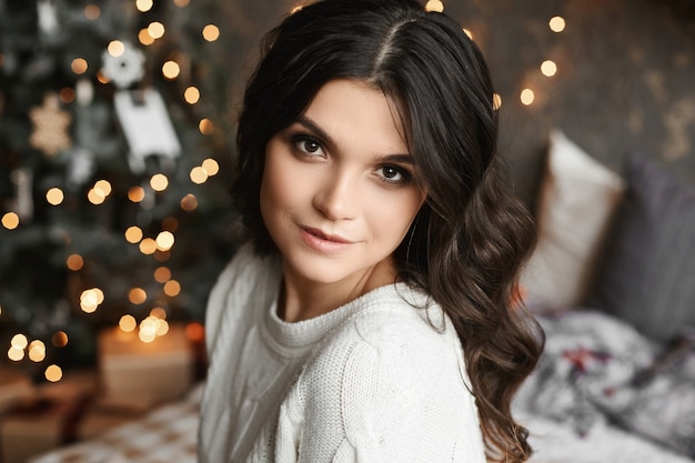 Portrait d'une jeune femme souriante posant dans un intérieur avec des lumières de Noël festives sur fond.