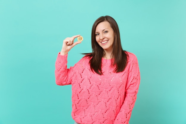 Portrait de jeune femme souriante en pull rose tricoté tenant à la main bitcoin, future monnaie isolée sur fond de mur turquoise bleu en studio. Concept de mode de vie des gens. Maquette de l'espace de copie.