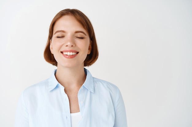 Portrait de jeune femme souriante rêveuse, fermer les yeux et rêver, debout en blouse contre le mur blanc