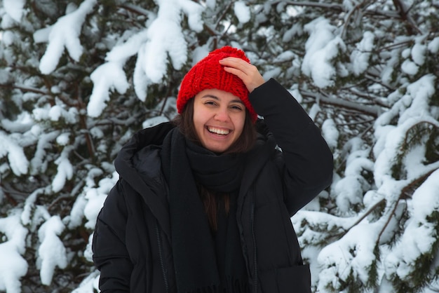 Portrait de jeune femme souriante en vêtements d'hiver au chapeau rouge avec sapin bubon en arrière-plan