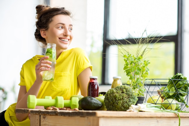 Portrait d'une jeune femme sportive en t-shirt jaune assise à l'intérieur avec des aliments sains et des haltères sur la table