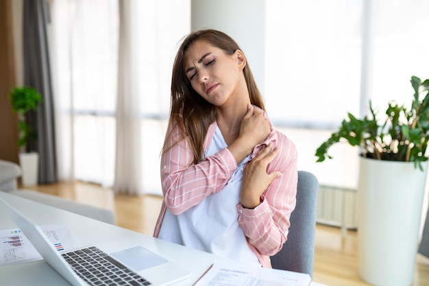 Portrait d'une jeune femme stressée assise au bureau à domicile devant un ordinateur portable touchant l'épaule douloureuse avec une expression douloureuse souffrant de mal à l'épaule après avoir travaillé sur un ordinateur portable