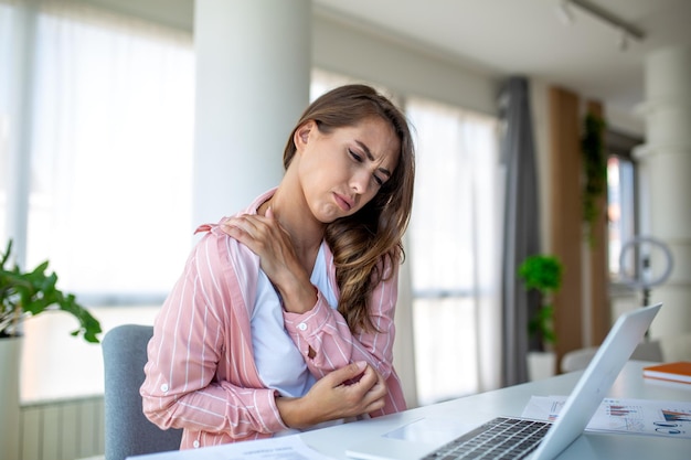 Portrait d'une jeune femme stressée assise au bureau du bureau à la maison devant un ordinateur portable touchant l'épaule douloureuse avec une expression de douleur souffrant d'une douleur à l' épaule après avoir travaillé sur un ordinateur laptop