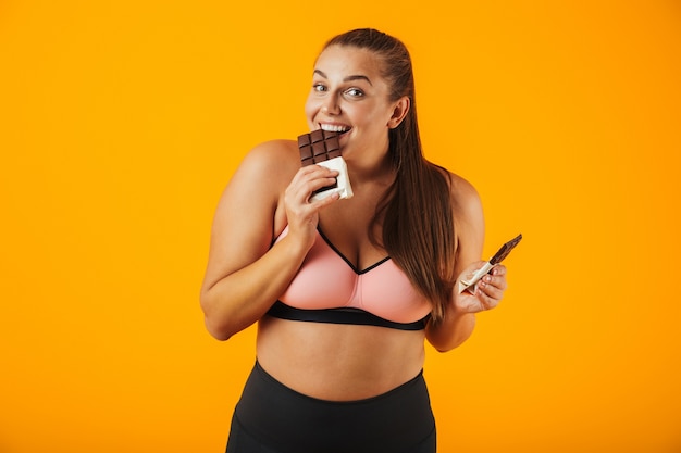 Portrait d'une jeune femme en surpoids ravie portant des vêtements de sport debout isolé sur mur jaune, manger du chocolat