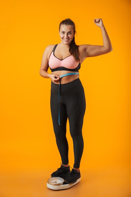 Portrait d'une jeune femme en surpoids satisfaits portant des vêtements de sport debout sur des échelles isolées sur un mur jaune, tenant un ruban à mesurer autour de sa taille