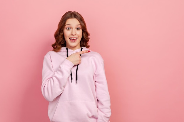 Portrait d'une jeune femme surprise aux cheveux bouclés en sweat à capuche regardant la caméra avec une expression étonnée heureuse pointant le doigt vers la droite sur l'espace de copie Studio intérieur tourné isolé sur fond rose