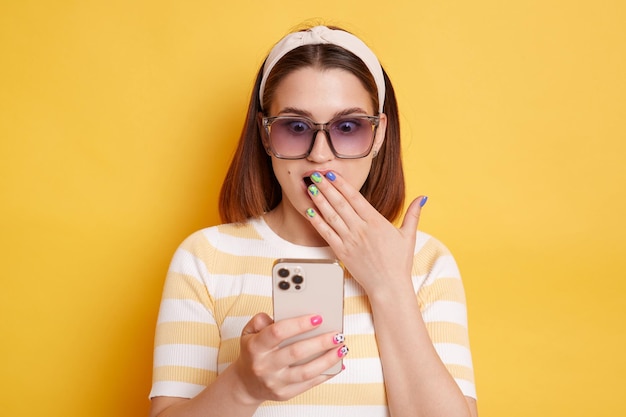 Portrait d'une jeune femme surprise choquée portant un t-shirt rayé tenant un téléphone portable lisant des nouvelles choquantes couvrant la bouche avec la paume posant isolé sur fond jaune