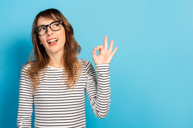 Portrait d'une jeune femme sympathique avec un sourire dans un t-shirt décontracté et des lunettes fait un geste correct et lève les yeux sur le bleu. Visage émotionnel. Le geste va bien, c'est OK