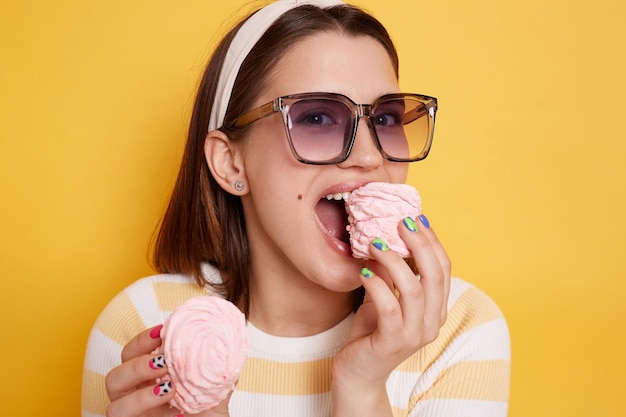 Portrait de jeune femme tenant des guimauves dans les mains portant une chemise rayée et des lunettes mangeant un dessert savoureux mordant des bonbons sucrés posant isolé sur fond jaune