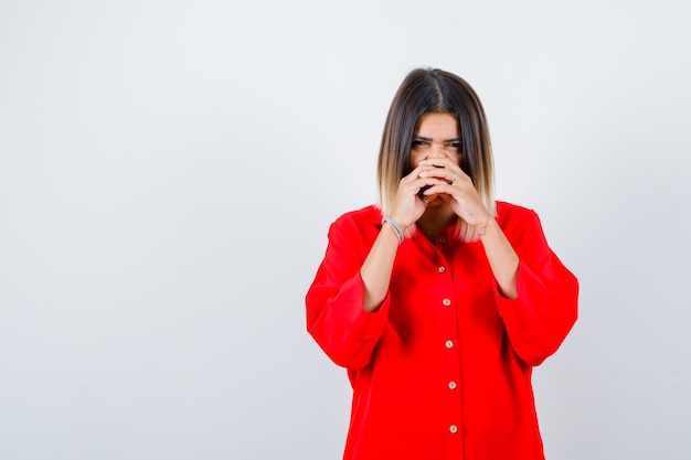 Portrait de jeune femme tenant les mains entrelacées sur le nez en chemise rouge surdimensionnée et à la jolie vue de face