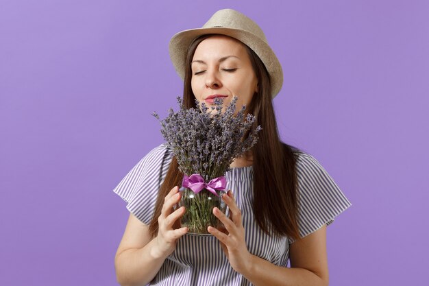 Portrait d'une jeune femme tendre et heureuse en robe bleue, chapeau de paille, tenir et renifler le bouquet de belles fleurs de lavande violettes isolées sur fond violet clair. Concept de vacances de la Journée internationale de la femme