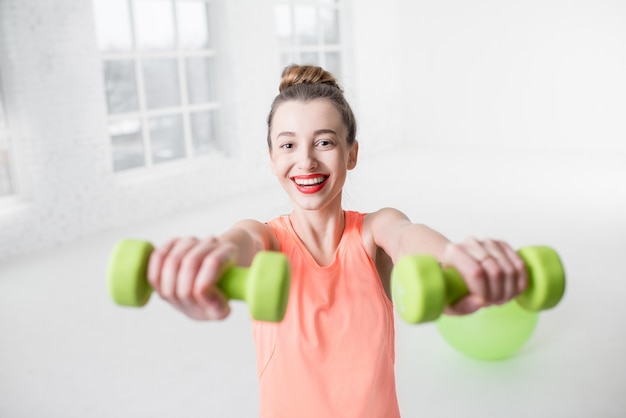 Portrait de jeune femme en tenue de sport avec haltères et ballon de fitness dans la salle de sport blanche