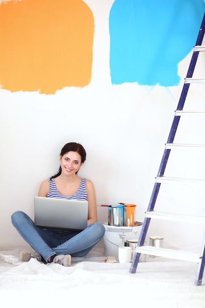 Portrait de jeune femme tout en peignant un nouvel appartement assis avec un ordinateur portable