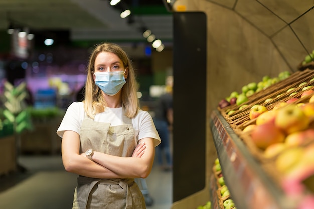 Portrait jeune femme travailleuse vendeuse dans un supermarché de la section Légumes debout dans un masque facial protégé les bras croisés. Marchande de fruits et légumes en regardant la caméra dans un marché de fruits Employé dans un tablier de travail