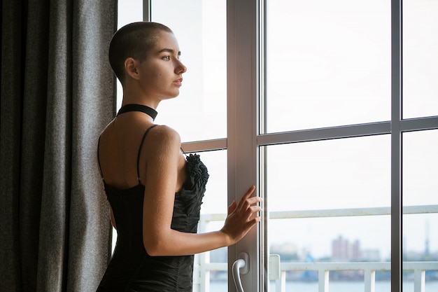 Portrait d'une jeune femme triste aux cheveux courts, regardant par la fenêtre dans une robe noire