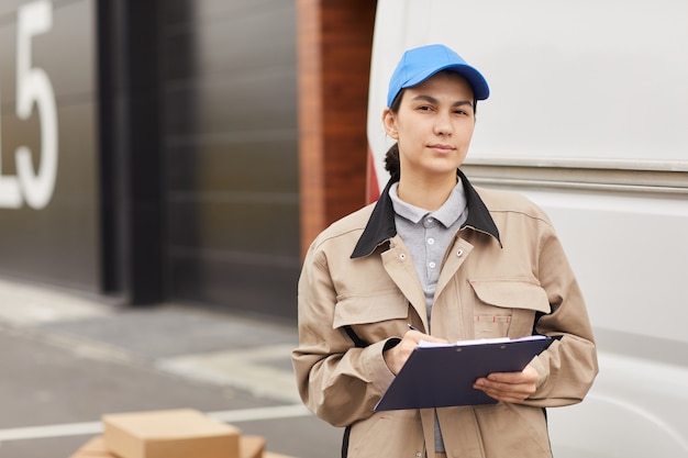 Portrait de jeune femme en uniforme remplissant le formulaire en se tenant debout à l'extérieur, elle travaille dans l'entrepôt de livraison