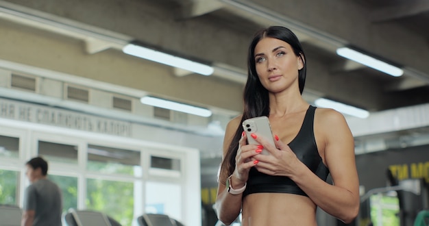 Portrait de jeune femme utilisant un smartphone au gymnase pendant l'entraînement. Message SMS de fille sportive souriante dans un club de sport. Femme se préparant à l'entraînement à l'aide de l'application de remise en forme.