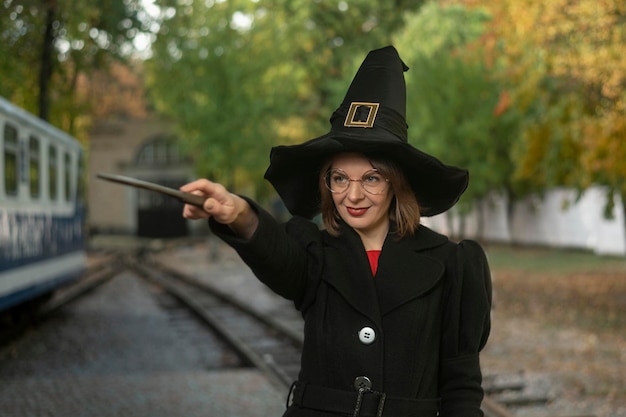 Portrait jeune femme vêtue d'un chapeau de sorcière, d'un manteau noir et de lunettes Fille souriante avec une baguette magique dans les mains Cadre vertical