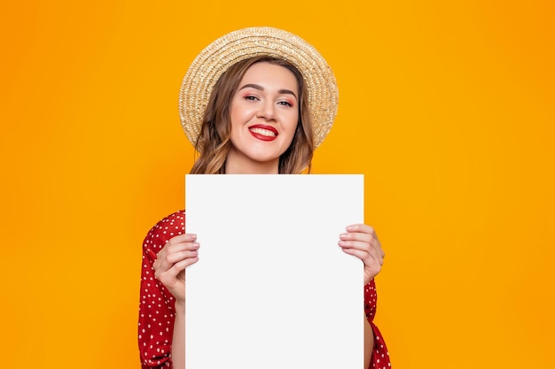 Portrait d'une jeune femme vêtue d'une robe rouge, d'un chapeau de paille aux lèvres rouges souriant et tenant dans ses mains une affiche au format A3 isolée sur fond orange, maquette