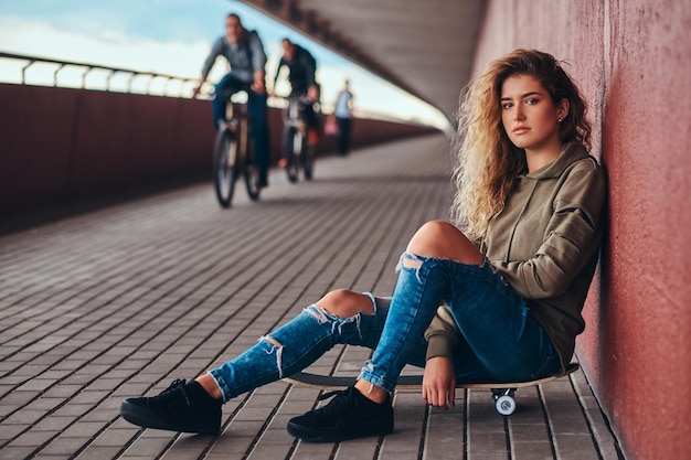 Portrait d'une jeune femme vêtue d'un sweat à capuche et d'un jean déchiré, appuyée sur un mur, assise sur une planche à roulettes au pied du pont.