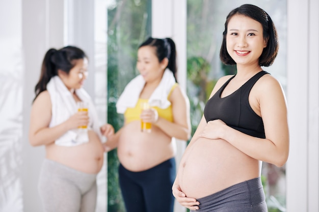 Portrait de jeune femme vietnamienne enceinte souriante touchant son ventre, ses amis enceintes parler en arrière-plan