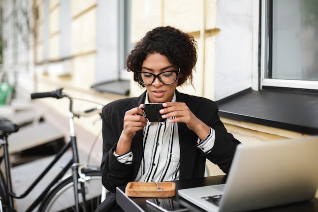 Portrait de jeune fille afro-américaine dans des verres assis à la table du café avec ordinateur portable et tenant une tasse de café dans les mains