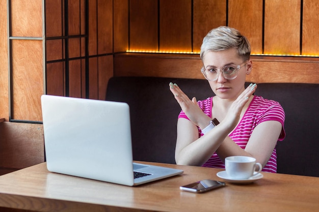 Portrait d'une jeune fille agressive avertissante aux cheveux courts blonds en t-shirt rose est assise dans un café et travaille sur un ordinateur portable et montre des bras levés croisés comme un geste d'arrêt Intérieur regardant la caméra