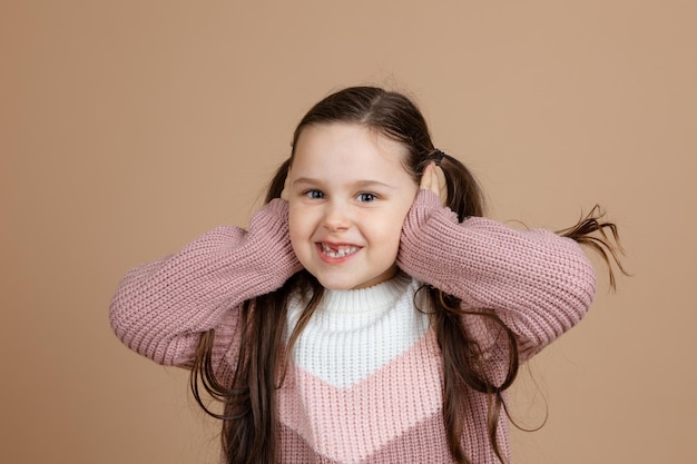 Portrait de jeune fille assez souriante géniale avec de longs cheveux noirs en pull rose blanc debout couvrant les oreilles avec les mains posant