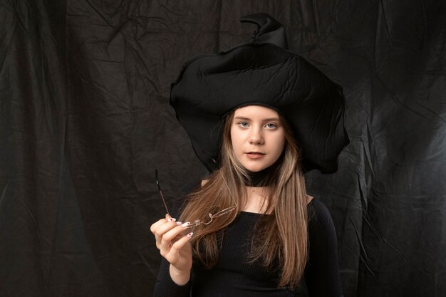 Portrait de jeune fille au chapeau de sorcière pointu avec des lunettes dans ses mains sur fond noir costume d'Halloween