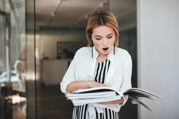 Portrait de jeune fille aux cheveux blonds tenant des livres dans les mains et regardant avec étonnement dessus. Fille choquée en robe dépouillée et chemise blanche debout près de la porte avec des journaux dans les mains