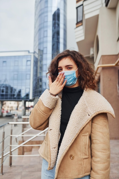 Portrait d'une jeune fille aux cheveux bouclés portant un masque de protection qui tousse à l'extérieur près d'un bâtiment commercial au moment de la quarantaine Conception du coronavirus