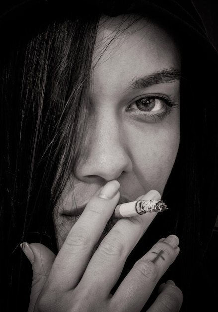 Portrait d'une jeune fille aux cheveux longs dans une cagoule qui tient dans ses doigts et fume une cigarette