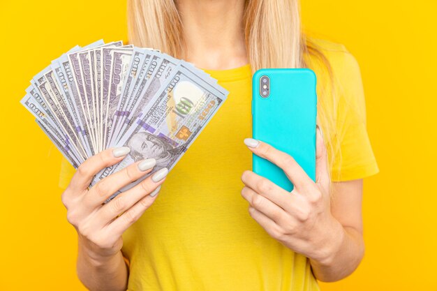 Portrait jeune fille aux longs cheveux blonds debout sur jaune, tenant des billets d'argent, à l'aide de téléphone mobile.