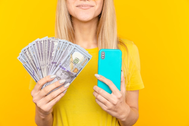 Portrait jeune fille aux longs cheveux blonds debout sur jaune, tenant des billets d'argent, à l'aide de téléphone mobile.