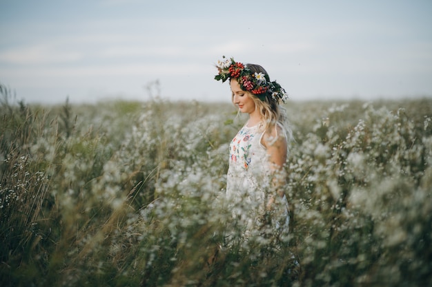 Portrait de jeune fille blonde aux yeux bleus avec une couronne de fleurs sur la tête marchant dans le champ avec des fleurs blanches