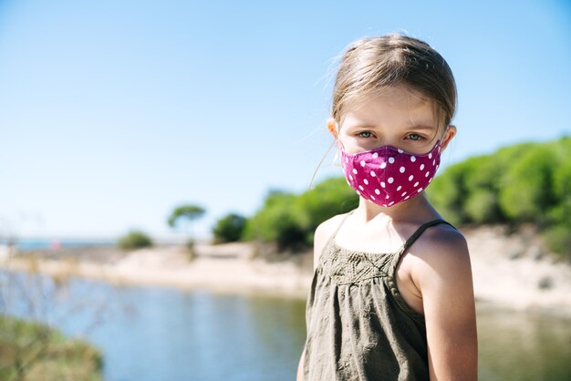 Portrait d'une jeune fille blonde aux yeux bleus portant un masque facial en vacances sur une plage de pins avec un visage sérieux dans une robe verte au milieu d'une pandémie de coronavirus