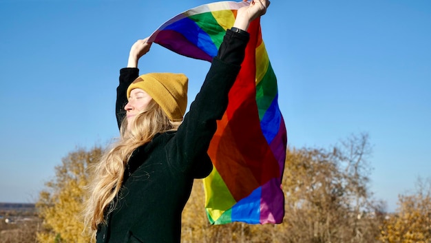 Portrait d'une jeune fille blonde caucasienne avec de l'acné tenant un drapeau LGBT arc-en-ciel en automne Femme au chapeau jaune et manteau noir sur une journée ensoleillée
