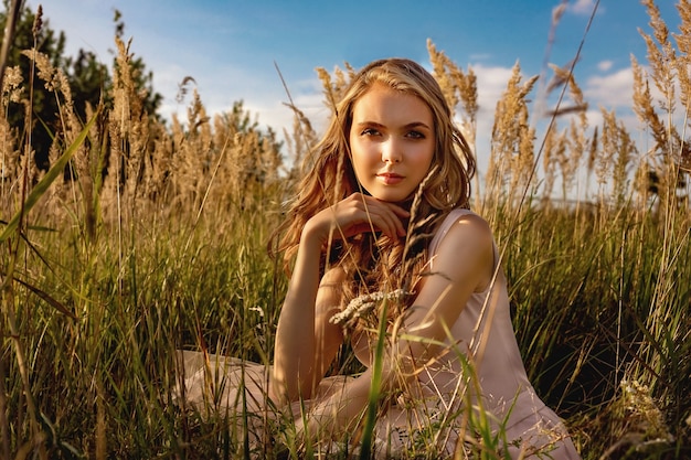 Portrait d'une jeune fille blonde dans une robe qui se trouve dans un champ avec de hautes herbes et un bouleau.