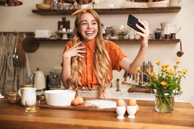 Portrait d'une jeune fille blonde émotionnelle joyeuse chef cuisinant à la cuisine prendre un selfie par téléphone portable.