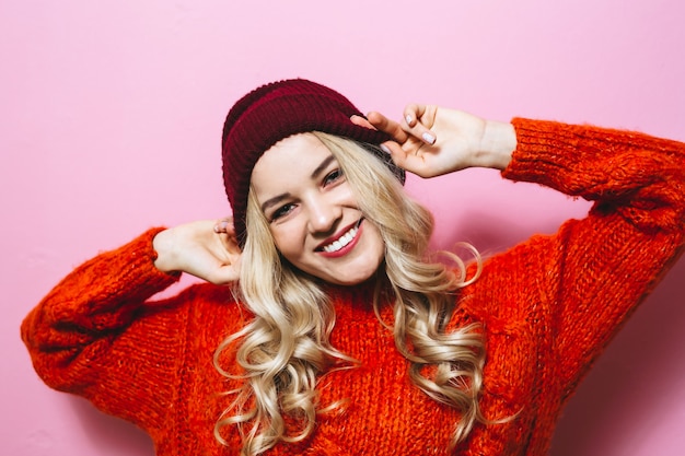 Portrait d'une jeune fille blonde portant une casquette et est vêtu d'un pull rouge et montrant la mode se déplace sur le mur rose