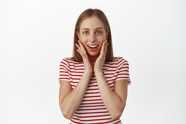 Portrait d'une jeune fille blonde souriante ravie se sent flattée et touchée, l'air surprise du cadeau, se tient la main près du visage et regarde la caméra avec admiration, debout sur fond blanc